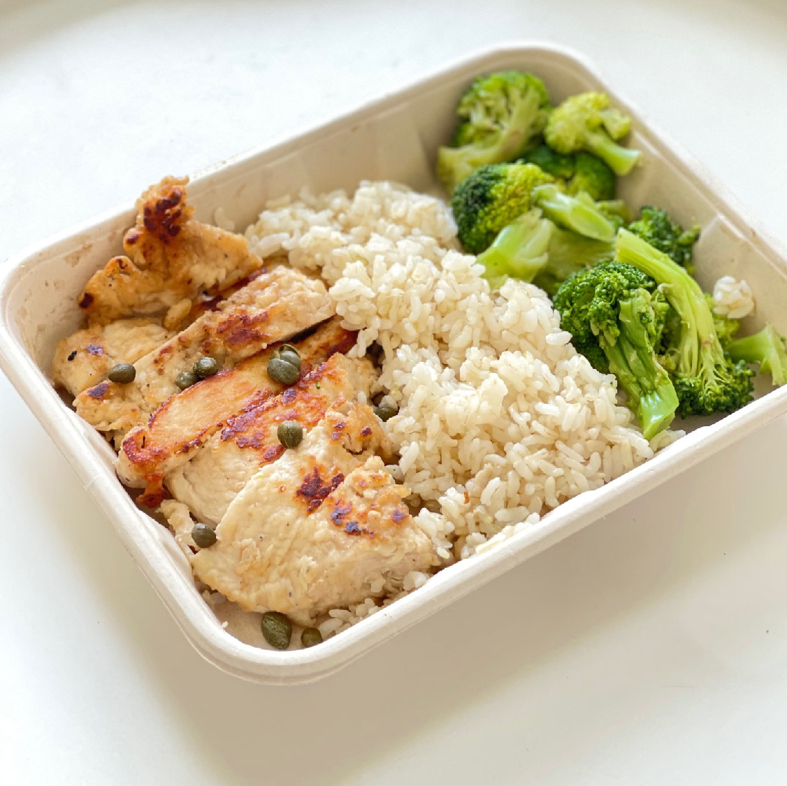 A photo of grilled chicken piccata with rice and steamed broccoli in a tan paper container..